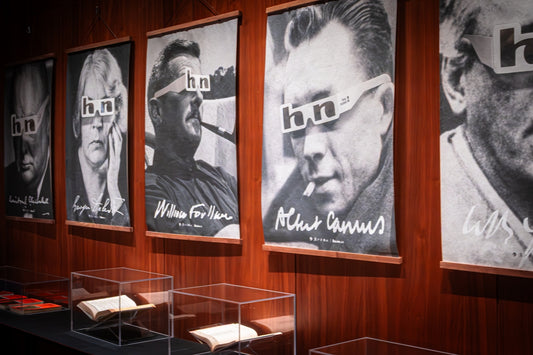 Portraits wearing 'hn' glasses displayed above books in a gallery.